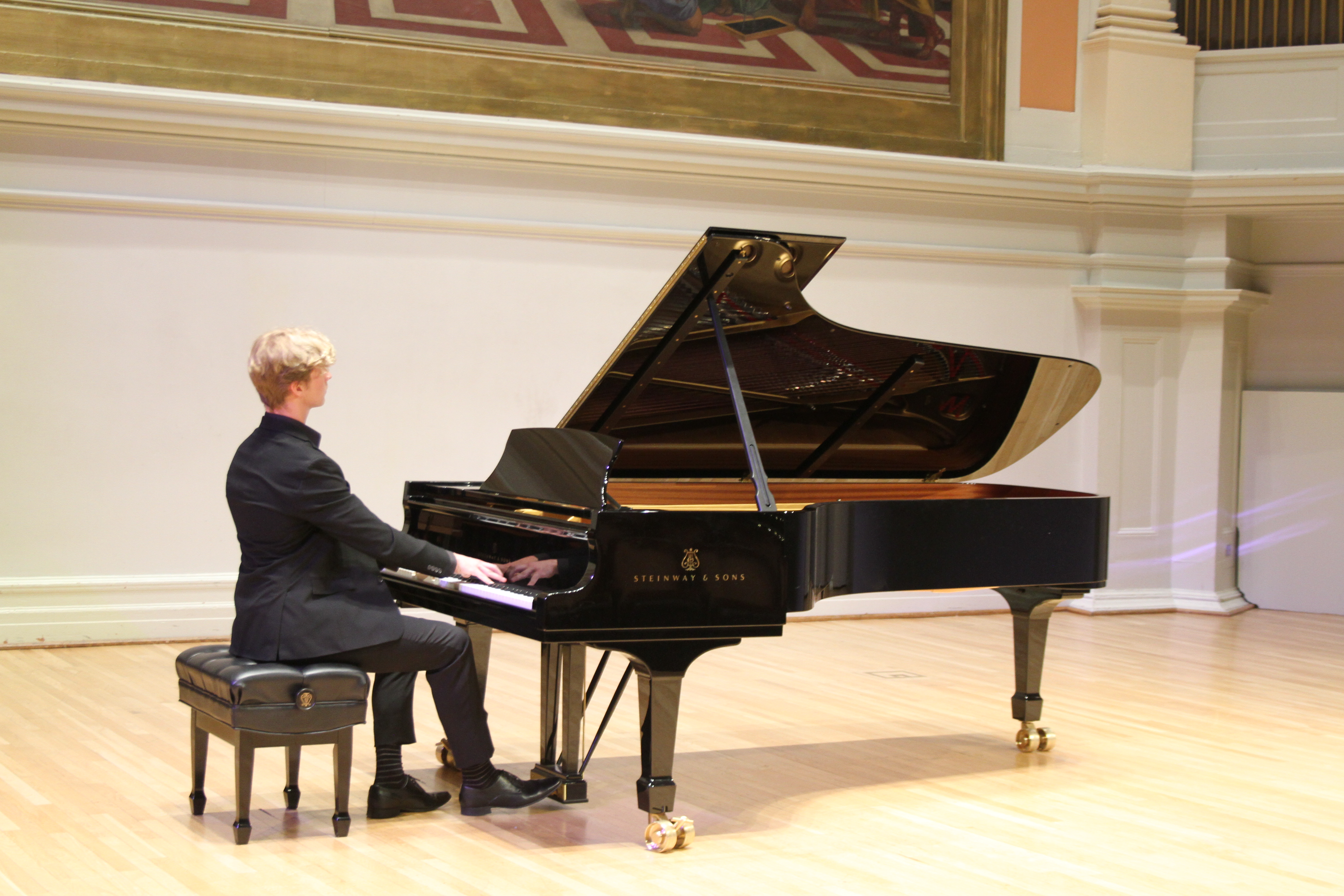 A photograph of a young man playing a grand piano on the stage of Old Cabell Hall