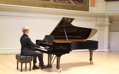 A photograph of a young man playing a grand piano on the stage of Old Cabell Hall